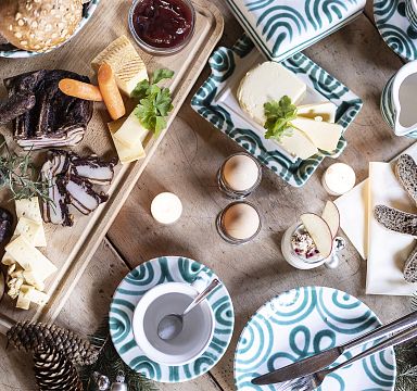 Eine gedeckte Frühstückstafel mit Brot, Käse, Aufschnitt und Eiern, dekoriert mit Kerzen, Tannenzapfen und weihnachtlichen Elementen auf einem Holztisch.