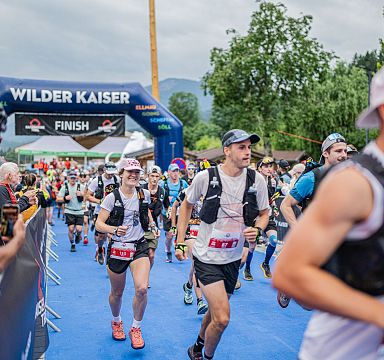 Teilnehmer eines Laufs überqueren die Ziellinie bei einem Event in der Bergregion Wilder Kaiser. Im Hintergrund sieht man Berge und Bäume.