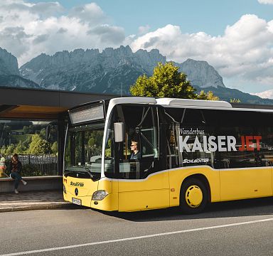 A yellow bus at a stop with people walking nearby, set against the backdrop of the Wilder Kaiser mountains and a partly cloudy sky.