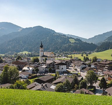 Ein malerisches Dorf in einer grünen Alpenlandschaft mit einer Kirche im Zentrum, umgeben von traditionellen Häusern, Wiesen und bewaldeten Bergen.