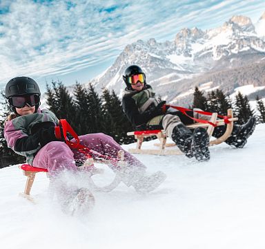 Zwei Personen rodeln fröhlich einen verschneiten Hang hinunter, umgeben von Nadelbäumen und einem beeindruckenden Bergmassiv im Hintergrund.
