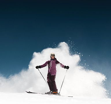 Eine Person in Skiausrüstung fährt dynamisch einen verschneiten Hang hinunter, umgeben von aufwirbelndem Schnee vor klarem, blauem Himmel.