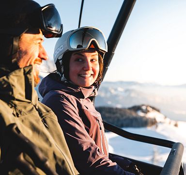 Zwei Skifahrer in Winterkleidung im Sessellift, umgeben von schneebedeckten Bergen und blauem Himmel. Sie lachen und genießen die winterliche Landschaft.