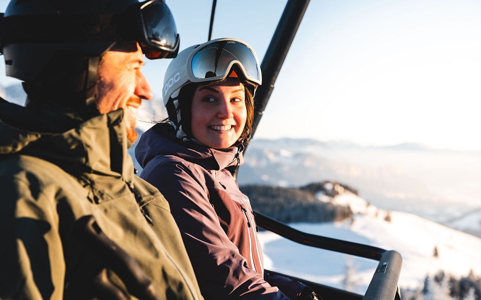 Zwei Skifahrer in Winterkleidung im Sessellift, umgeben von schneebedeckten Bergen und blauem Himmel. Sie lachen und genießen die winterliche Landschaft.