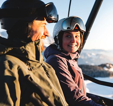 Zwei Skifahrer in Winterkleidung im Sessellift, umgeben von schneebedeckten Bergen und blauem Himmel. Sie lachen und genießen die winterliche Landschaft.