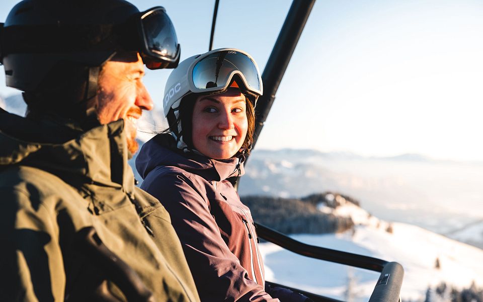 Zwei Skifahrer in Winterkleidung im Sessellift, umgeben von schneebedeckten Bergen und blauem Himmel. Sie lachen und genießen die winterliche Landschaft.