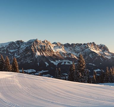 Verschneite Skipiste mit perfekt präparierten Spuren, im Hintergrund das beeindruckende Bergmassiv des Wilden Kaisers bei klarem Himmel und Sonnenuntergang.