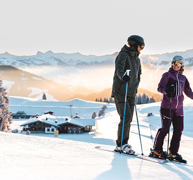 Zwei Skifahrer stehen auf einer verschneiten Piste mit Blick auf Berggipfel im Hintergrund. Die Sonne geht auf und schafft eine beeindruckende Kulisse.