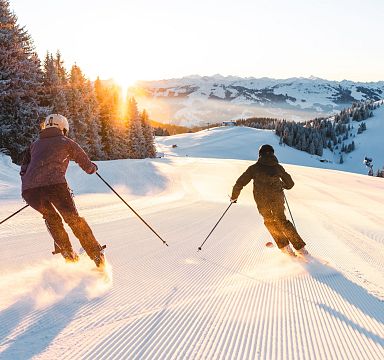 Zwei Skifahrer fahren bei Sonnenaufgang eine schneebedeckte Piste hinunter, umgeben von verschneiten Bäumen und Bergen im Hintergrund.