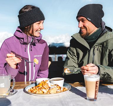 Zwei Personen in Winterkleidung genießen auf einer Hütte im Freien ein Dessert und Heißgetränke. Schnee bedeckt die Umgebung, sonnig, klare Himmel.