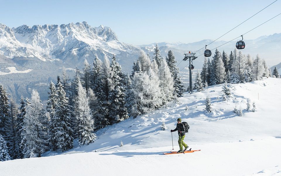 Ein Skifahrer fährt einen schneebedeckten Hang hinunter, umgeben von verschneiten Bäumen. Im Hintergrund sind Seilbahnkabinen und ein beeindruckendes Bergpanorama zu sehen.