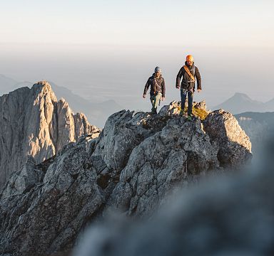 Two hikers on a rugged, rocky ridge with the dramatic peaks of the Wilder Kaiser mountains in the background, under a clear sky.