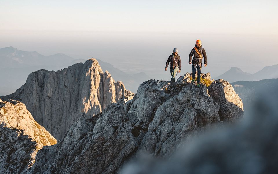 Zwei Wanderer stehen auf einem felsigen Berggipfel mit beeindruckender Aussicht auf umliegende Täler und Berge bei Sonnenuntergang.