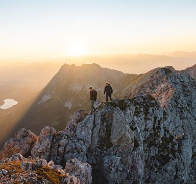 Zwei Wanderer stehen bei Sonnenaufgang auf einem felsigen Berggipfel. Im Hintergrund erheben sich weitere Berge und ein See ist in der Ferne sichtbar.