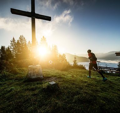 Ein Wanderer joggt bei Sonnenaufgang auf einem Berggipfel vorbei an einem großen Holzkreuz. Im Hintergrund sind Berge und Nebel im Tal zu sehen.