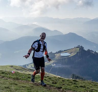 Ein Mann in Sportkleidung läuft auf einem Bergpfad mit malerischer Alpenlandschaft im Hintergrund, unter blauem Himmel und vorbei an sanften Hügelformen.