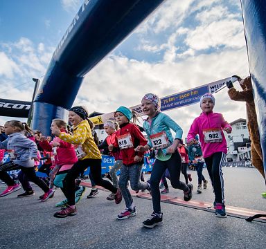 Eine Gruppe von Kindern läuft bei einem Rennen über die Ziellinie. Alle sind sportlich gekleidet, es herrscht eine dynamische und fröhliche Atmosphäre.