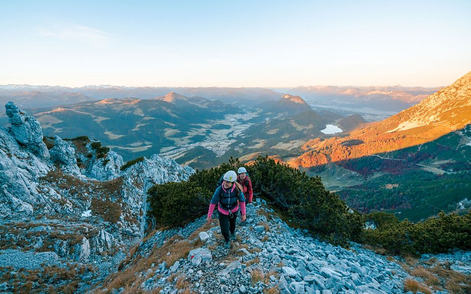Wanderer erklimmen einen steinigen Bergpfad bei Sonnenaufgang, umgeben von dramatischen Felsformationen und einem weiten Tal im Hintergrund.