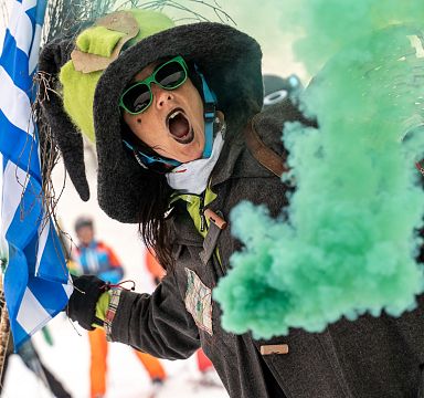 Person in Winterkleidung mit grünem Rauch und Fahne, in einer verschneiten Umgebung. Sie trägt Sonnenbrillen und einen markanten Hut.