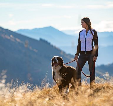 Frau in sportlicher Kleidung steht mit einem Hund auf einem Berg, umgeben von malerischen Bergen und blauem Himmel im Hintergrund an einem sonnigen Tag.
