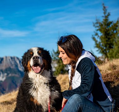 Frau sitzt lächelnd neben einem großen, flauschigen Hund in alpiner Landschaft mit Bergen im Hintergrund und blauem Himmel darüber. Herbstliches Setting.