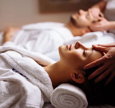 Zwei Personen in weißen Bademänteln genießen eine entspannende Kopfmassage in einem Spa. Sie liegen auf weichen Handtüchern und wirken entspannt und erholt.
