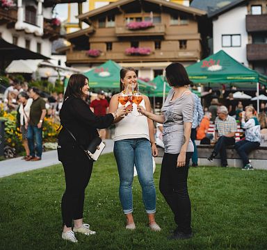 Drei Frauen stoßen mit Aperol Spritz Gläsern an, im Hintergrund traditionelle Tiroler Gebäude und Berglandschaft, sommerliche Atmosphäre.