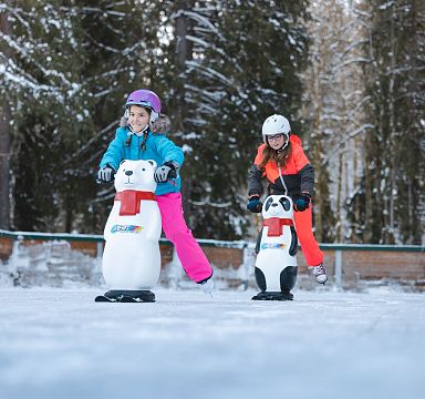 Zwei Kinder im Schnee mit Eislaufhilfen in Form von Tieren, umgeben von schneebedeckten Bäumen. Beide tragen bunte Winterkleidung und Helme.