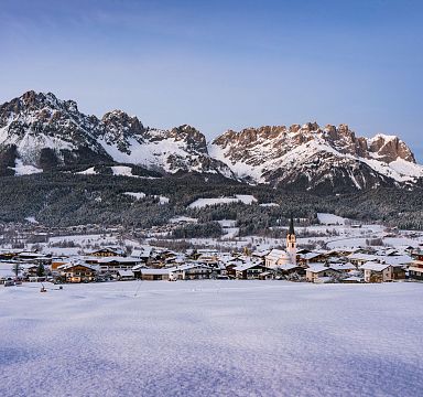 A picturesque winter scene with a snow-covered village below the rugged peaks of the Wilder Kaiser mountains, under a clear blue sky.