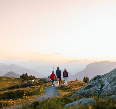 Three people hike a mountain path at sunset in the Wilder Kaiser, surrounded by rugged terrain and overlooking valleys. A cross stands on the trail ahead.