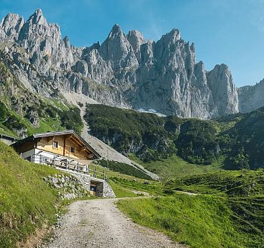Berglandschaft mit grünen Wiesen und einem holzverkleideten Haus im Vordergrund, dahinter ragen schroffe, felsige Gipfel unter einem blauen Himmel empor.