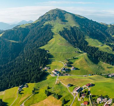 Panorama einer alpinen Landschaft mit grünen Hügeln und Wäldern. Kleine Dörfer und Straßen sind zu sehen, vor einem blauen Himmel unter leichtem Wolkenband.