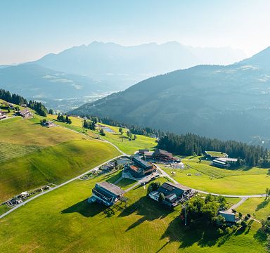 Aerial view of green hills and scattered houses beneath the towering Wilder Kaiser mountains in Tirol, Austria, on a clear day.