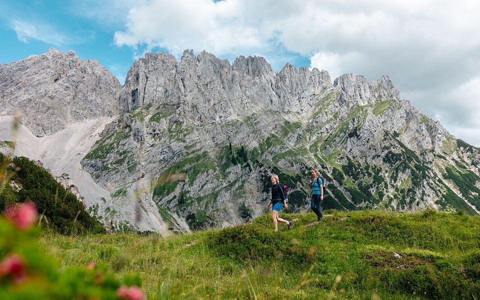 Two hikers walk through a green meadow with the jagged peaks of the Wilder Kaiser mountains in the background, under a partially cloudy sky.