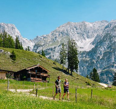 Two people hiking by a rustic alpine hut in lush green meadows with the majestic Wilder Kaiser mountains in the background under a clear blue sky.