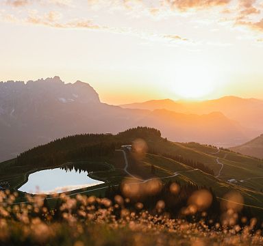 Sonnenuntergang über einer bergigen Landschaft mit einem kleinen See im Vordergrund und Silhouetten von Bergspitzen im Hintergrund.