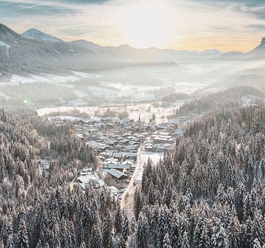 A serene winter scene at Wilder Kaiser with snow-covered trees and mountains framing a peaceful village under a warm sunrise.
