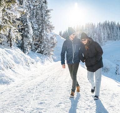 Zwei Menschen spazieren lachend auf einem verschneiten Weg in einer winterlichen Landschaft mit Sonnenschein, umgeben von schneebedeckten Bäumen.