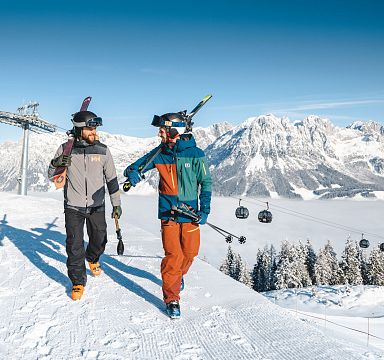 Two skiers walk near a chairlift in the Wilder Kaiser region, with snowy mountains in the background and a clear blue sky above, capturing winter sports excitement.