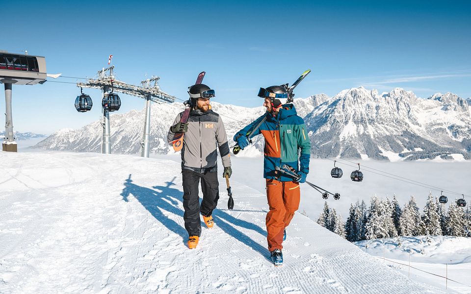 Zwei Skifahrer in winterlicher Berglandschaft am Wilder Kaiser tragen Ski auf den Schultern, während im Hintergrund Skilifte und verschneite Gipfel zu sehen sind.