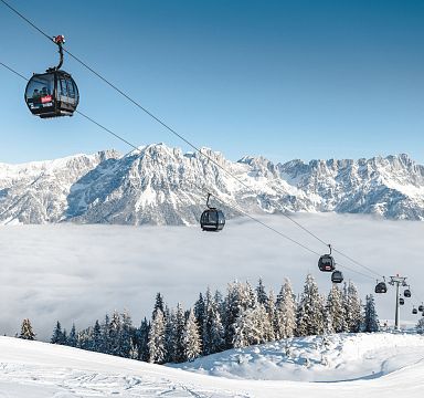 Schneebedeckte Berglandschaft mit Seilbahn und Gondeln über einer Wolkendecke, umgeben von verschneiten Bäumen und majestätischen Alpen im Hintergrund.