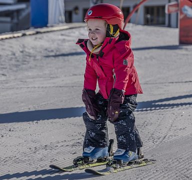 Ein kleines Kind in roter Jacke und rotem Helm fährt fröhlich Ski auf einer Piste. Im Hintergrund sind Schnee und Skiausrüstung zu sehen.