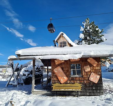 Snow-covered wooden cabin in the Wilder Kaiser region with a cable car above, set against a bright blue sky and snowy landscape.