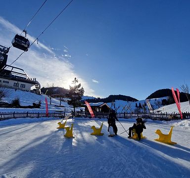 Winterlandschaft mit Sessellift, Kindern auf gelben Schlitten und verschneiten Bergen im Hintergrund, blauer Himmel und Sonnenschein.