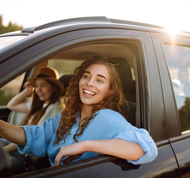Two people sitting in a parked car, enjoying the sunlight. The person in front is smiling and leaning out of the window, while the other sits in the passenger seat.