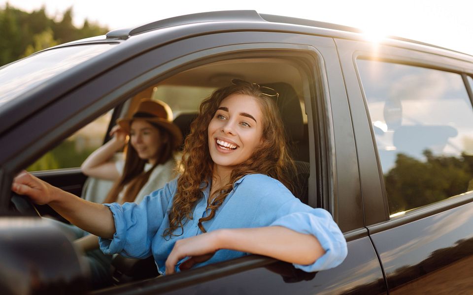 Zwei junge Frauen sitzen in einem Auto. Die Fahrerin lächelt entspannt, während die Sonne durch das Fenster scheint, und eine Passagierin sitzt im Hintergrund.