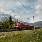 Roter Zug fährt durch grüne Landschaft, im Hintergrund beeindruckendes Bergmassiv unter blauem Himmel mit Wolken.