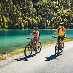 Zwei Radfahrer fahren auf einer Straße entlang eines klaren, türkisfarbenen Sees, umgeben von dicht bewaldeten Hügeln bei sonnigem Wetter.