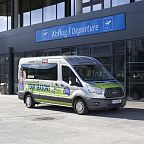 A shuttle van parked outside the departure terminal of an airport, displaying an advertisement on its side. The building has glass doors and a blue sign.