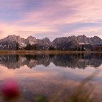 The Wilder Kaiser mountains reflect in a tranquil lake at sunset, with the sky painted in pink and purple tones, creating a serene and picturesque view.